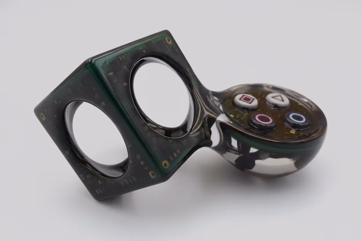 Fidget toy combining a transparent cube with circuit-like patterns and a rounded section with four PlayStation-style buttons.